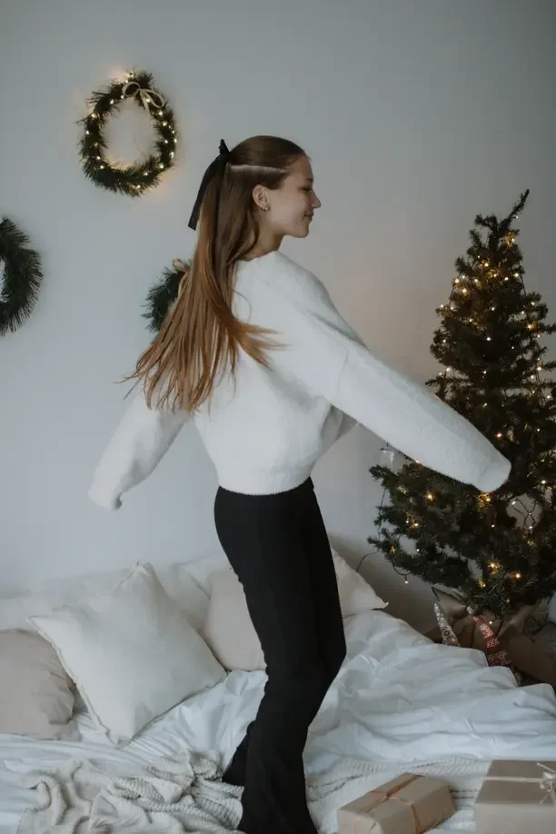 Christmas gifts for twenty somethings with a young woman in a white sweater twirling beside a Christmas tree.